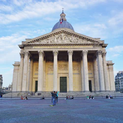 En neoklassisk byggnad med pelare och en kupol, människor sitter och går framför, med intilliggande stadsbyggnader under en blå himmel.