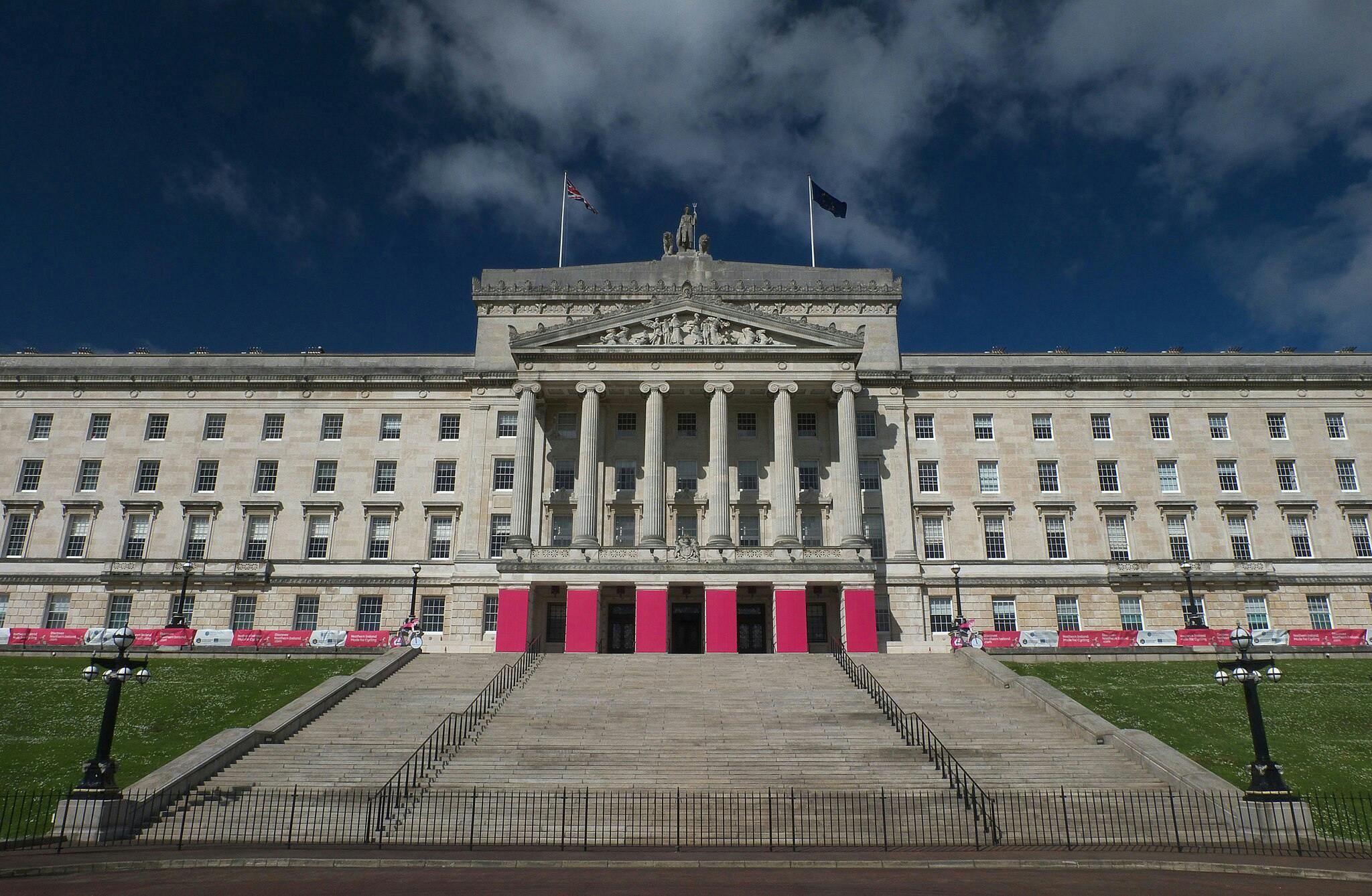 En stor neoklassisk byggnad med pelare, breda trappsteg, två flaggor på taket och rosa affischer nära ingångarna.