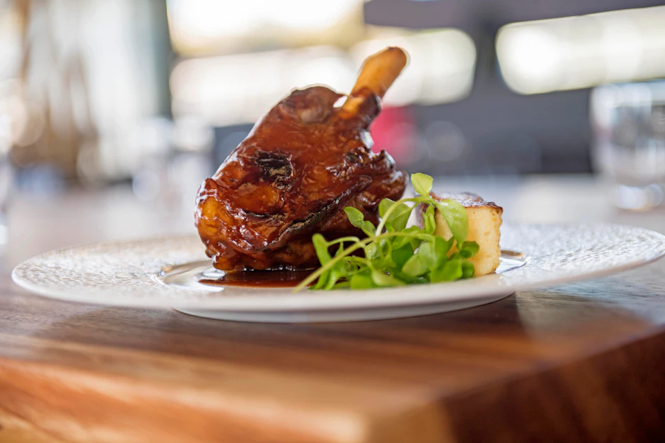 Plate with glazed meat, garnished with greens, and served with a side of mashed potatoes on a wooden board. Blurred background.