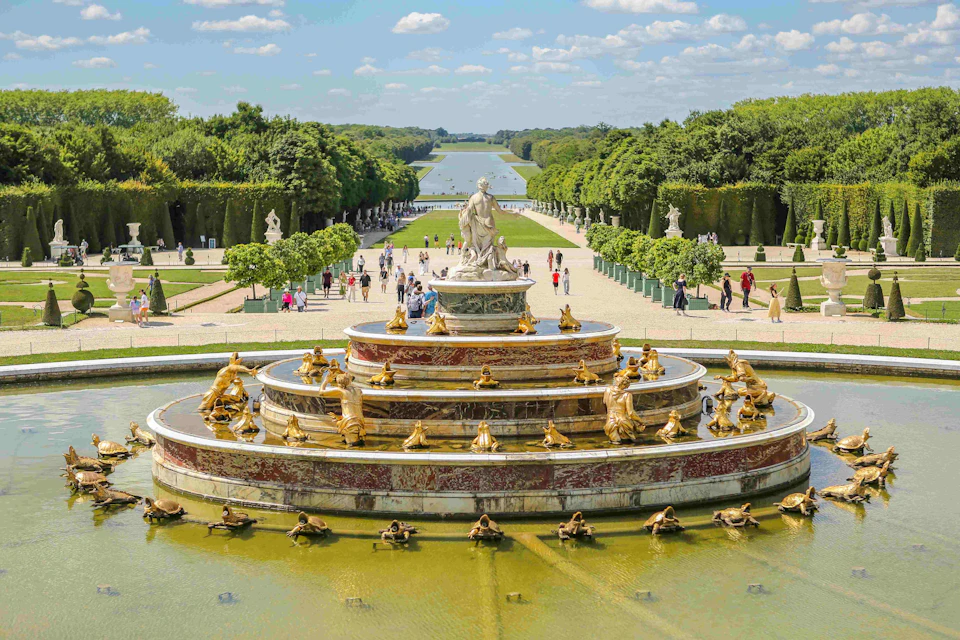 Ornate fountain with golden statues and tiers, surrounded by manicured gardens, pathways, and visitors under a partly cloudy sky.