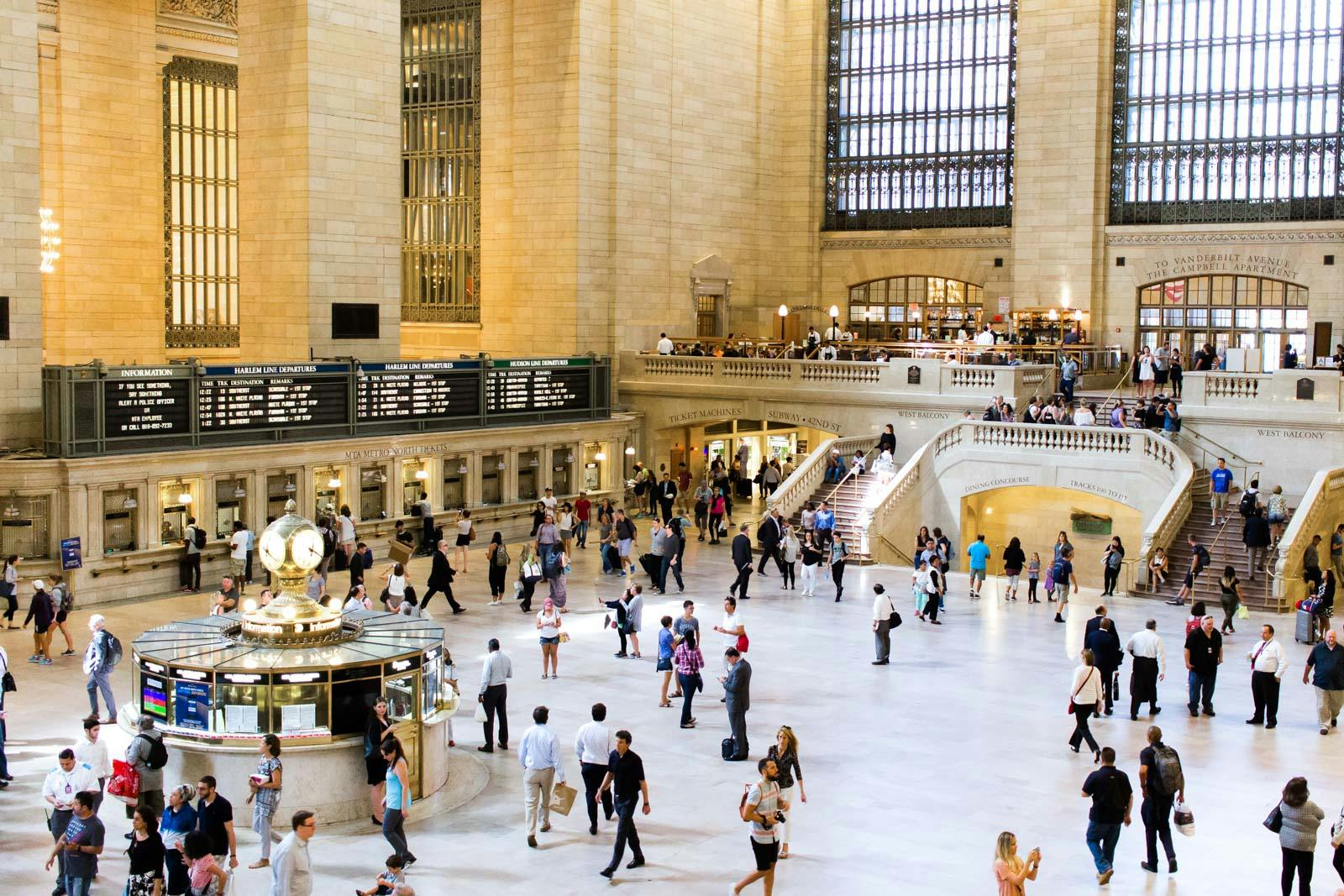 Busy terminal hall with people walking, a central clock, lit stairs, information boards, and large arched windows.