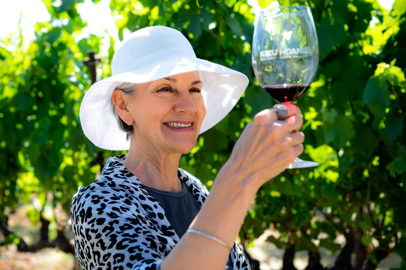 一位戴着太阳帽的微笑女士在绿色葡萄园藤蔓的背景下举起一杯红酒。