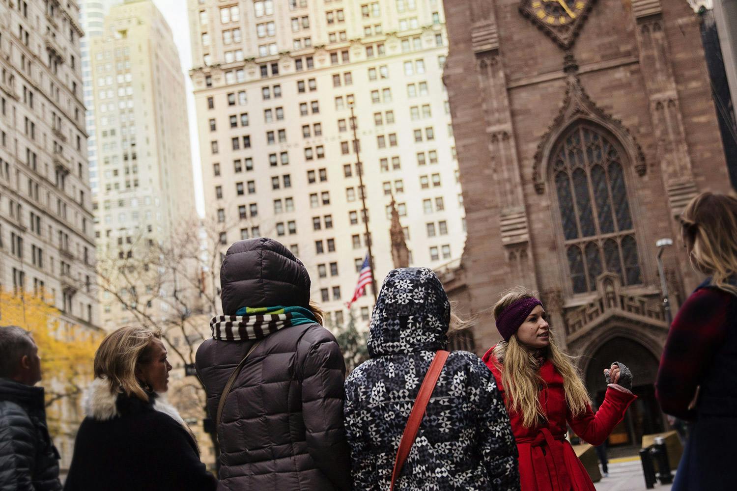 A woman in a red coat speaks to three other people outside a Gothic-style church in an urban setting.
