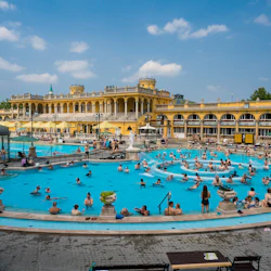 Människor simmar och kopplar av i flera stora pooler i en utsmyckad, historisk byggnad under en klarblå himmel.