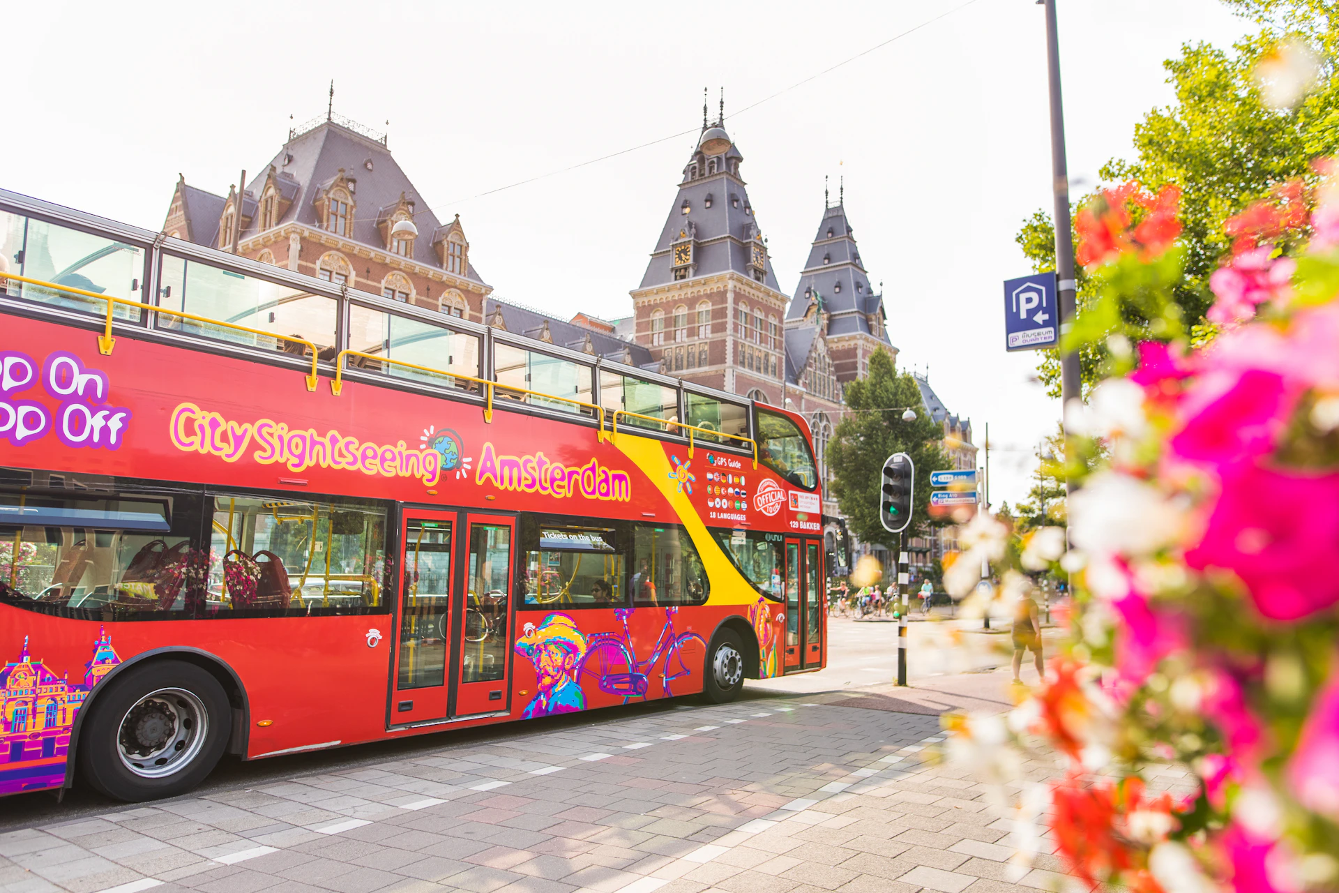A red double-decker sightseeing bus with "City Sightseeing Amsterdam" parked near historic buildings on a sunny day.