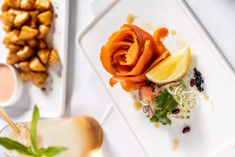 Elegant dish featuring a rose-shaped smoked salmon, a lemon wedge, sprouts, and garnishes on a white rectangular plate.