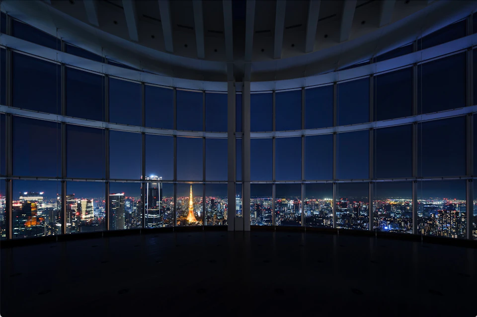 A nighttime cityscape seen through large curved windows, with a prominent lit tower in the center.