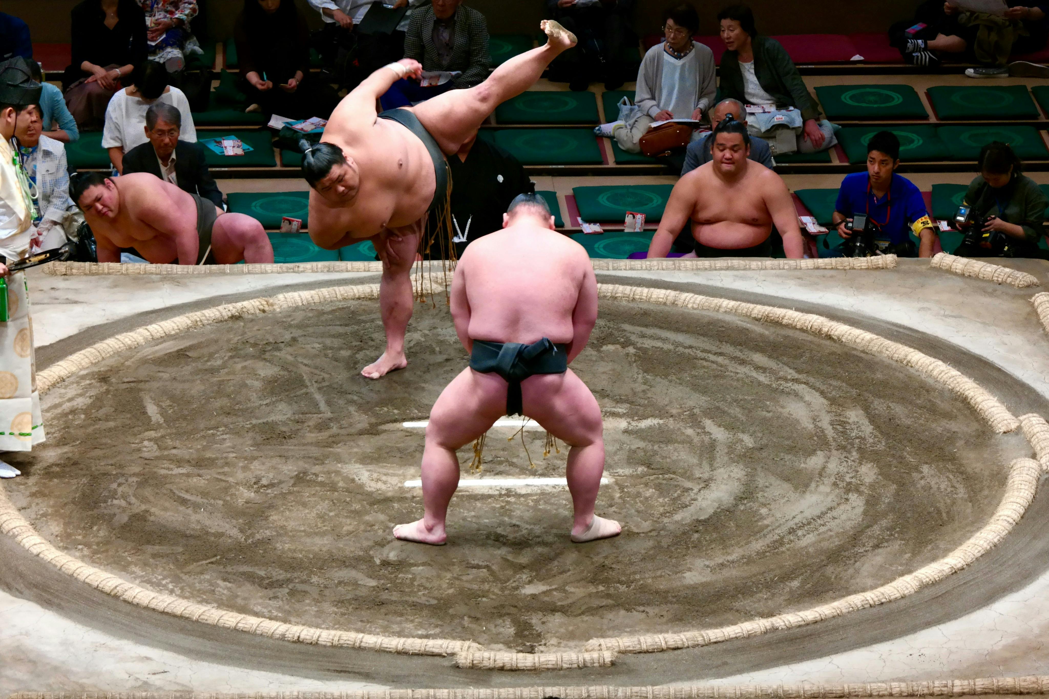Two sumo wrestlers in a match, one lifting a leg, on a circular ring surrounded by spectators and photographers.