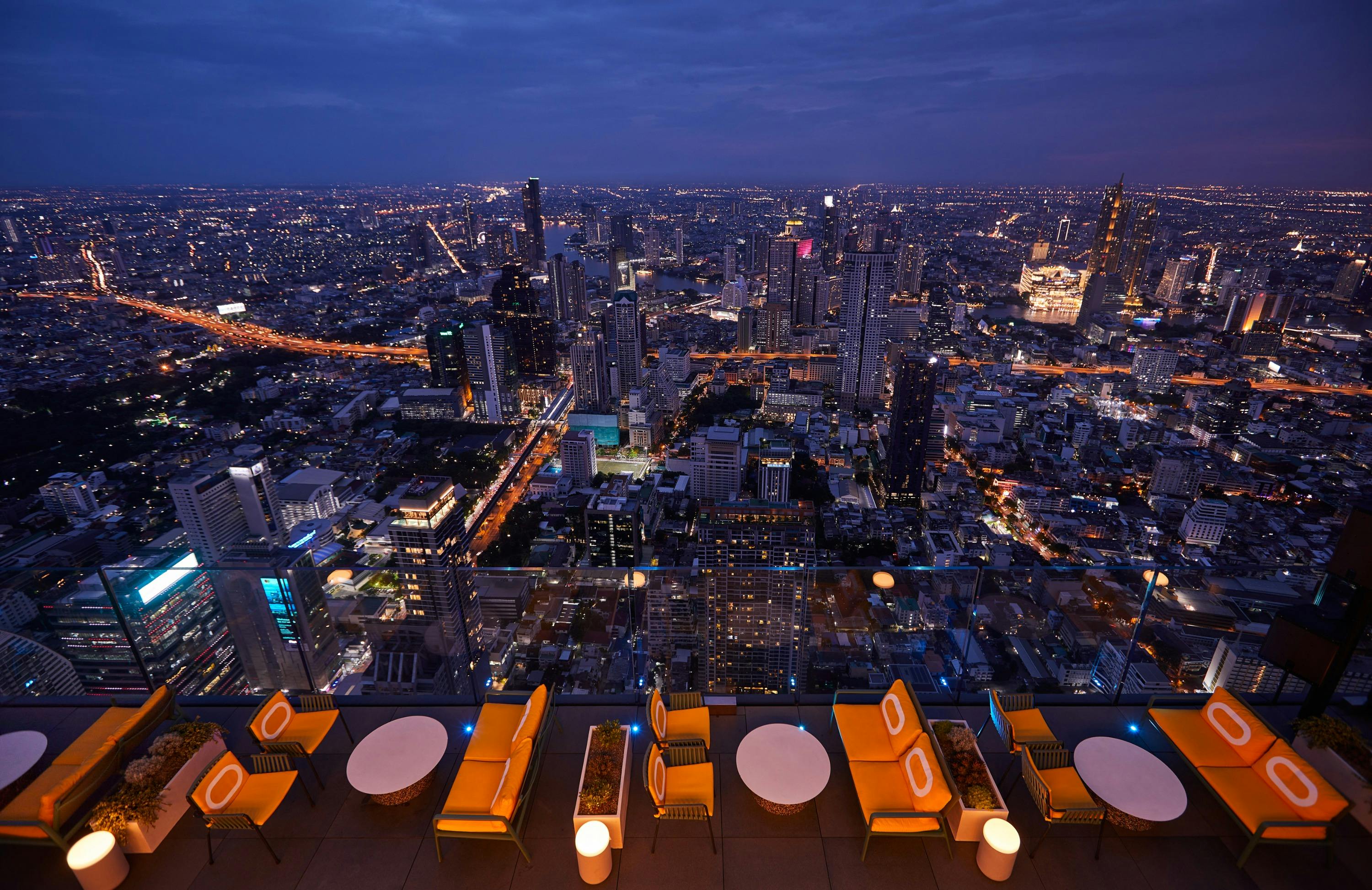 Rooftop restaurant with yellow chairs and round tables overlooking a brightly lit cityscape at night.