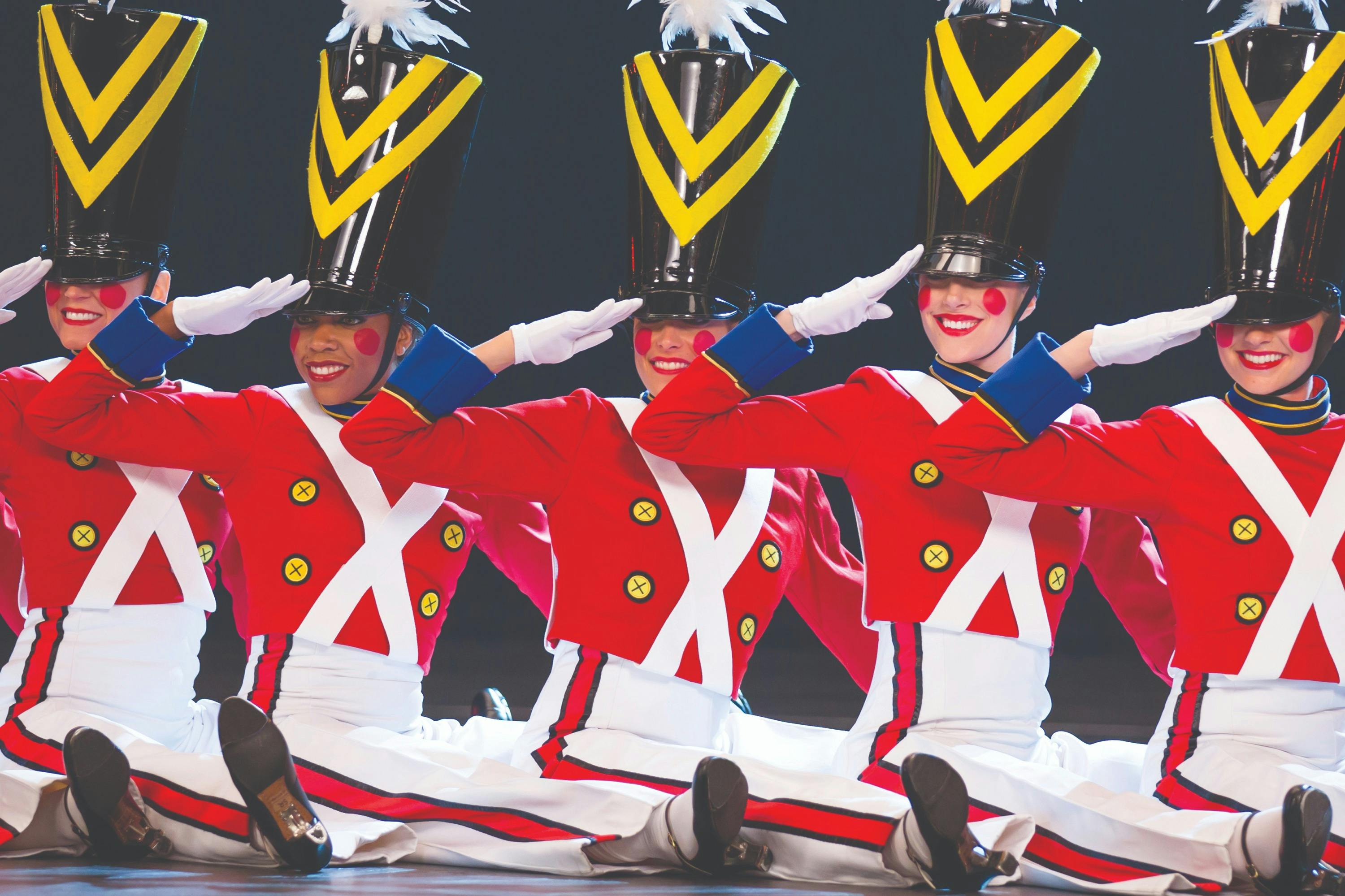 Um grupo de artistas vestidos de soldadinhos de chumbo, com uniformes vermelhos e brancos, saúda-os com sorrisos no palco.