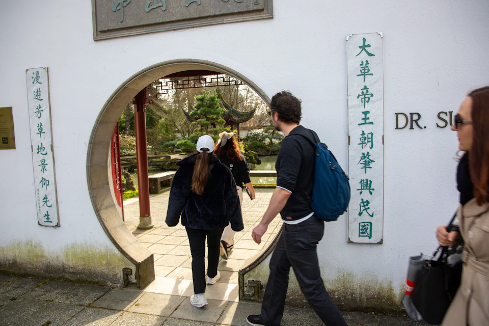 People walk through a circular entrance in a white wall with Chinese characters, leading to a garden with greenery and traditional architecture.
