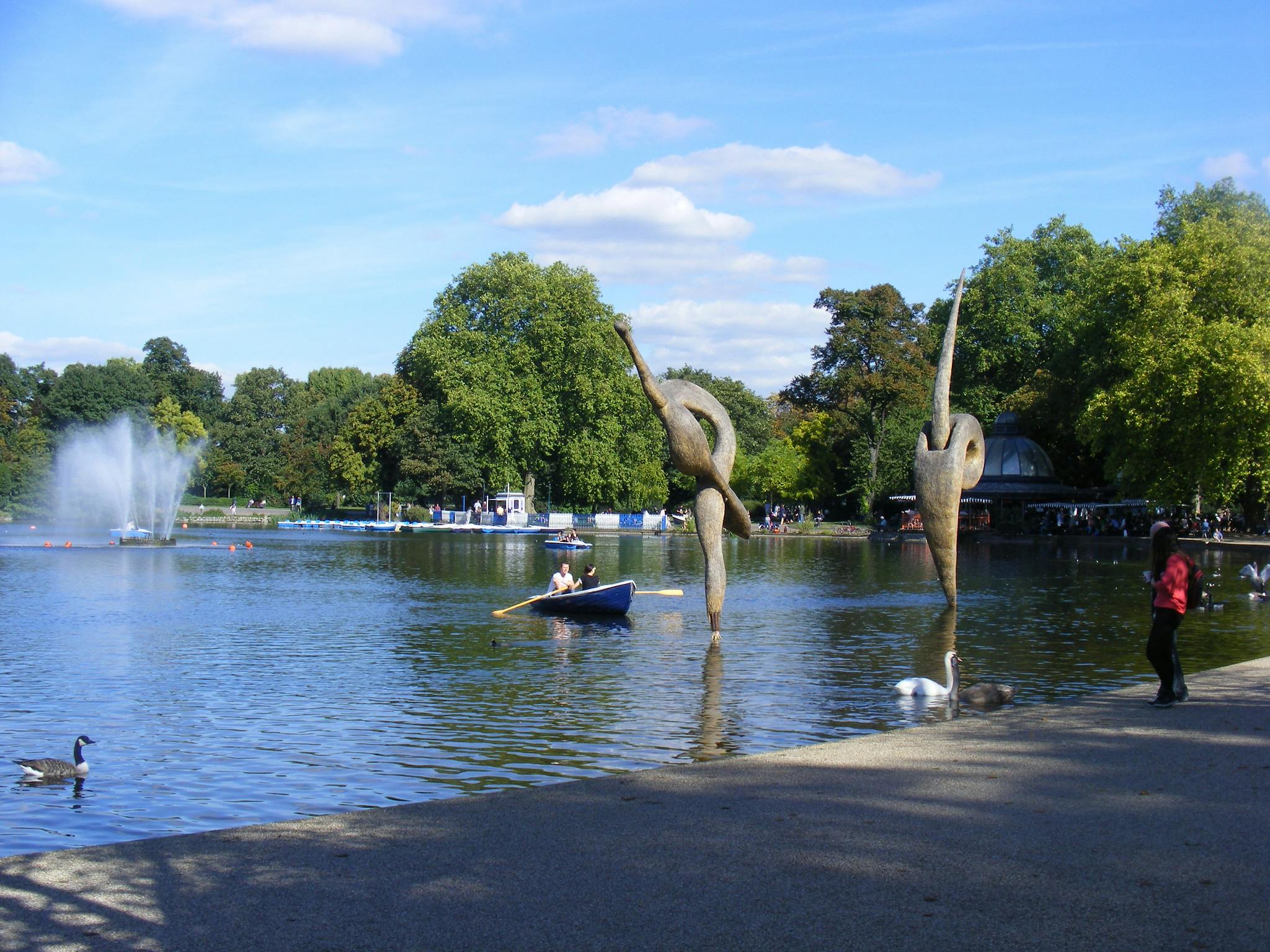 Сцена в парке с озером, фонтаном, скульптурами, гребной лодкой, лебедями, утками и деревьями под пасмурным небом.