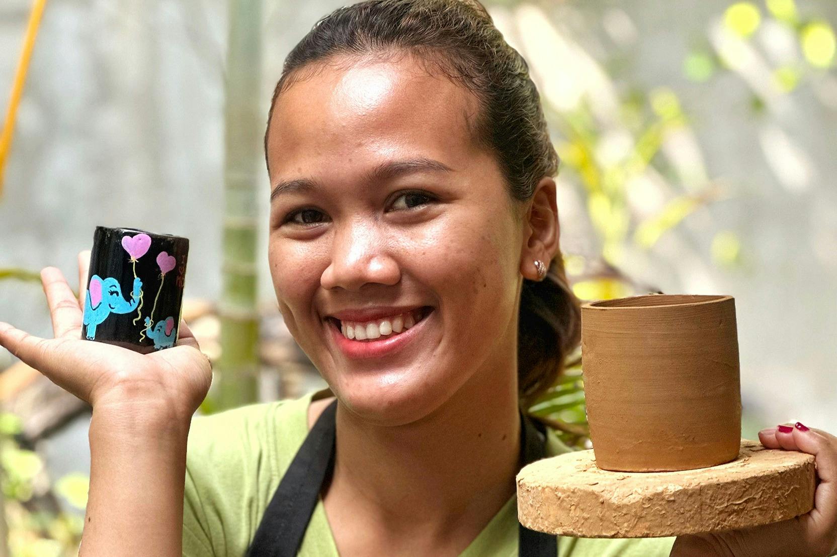 A smiling person holding a painted black pot with balloon designs, standing next to a plain terracotta pot outdoors.