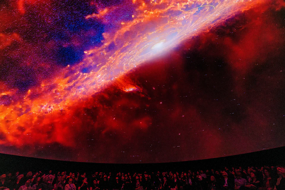 A crowd of people observing a vivid cosmic projection of a galaxy's center in a dark theater-like environment.
