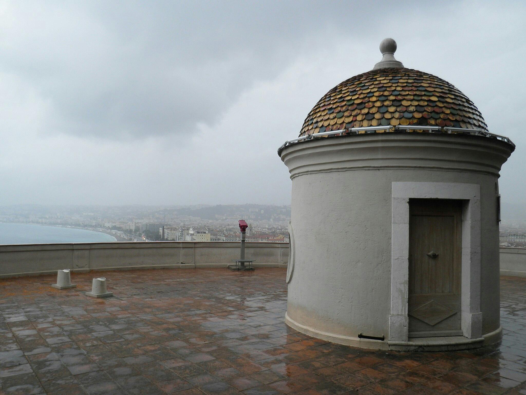 这栋小圆形建筑位于潮湿的露台上，配有彩色瓷砖圆顶，可俯瞰多云的天空下的城市景观和海岸线。
