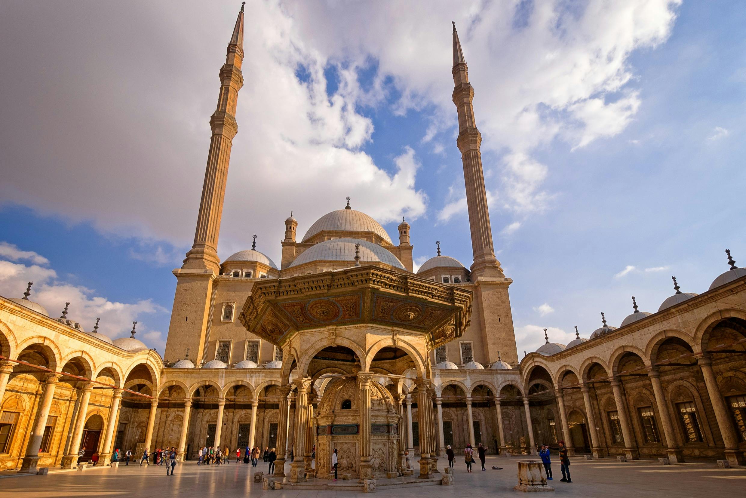 A large mosque courtyard with two tall minarets, a central ornate structure, domes, and a blue sky with clouds.