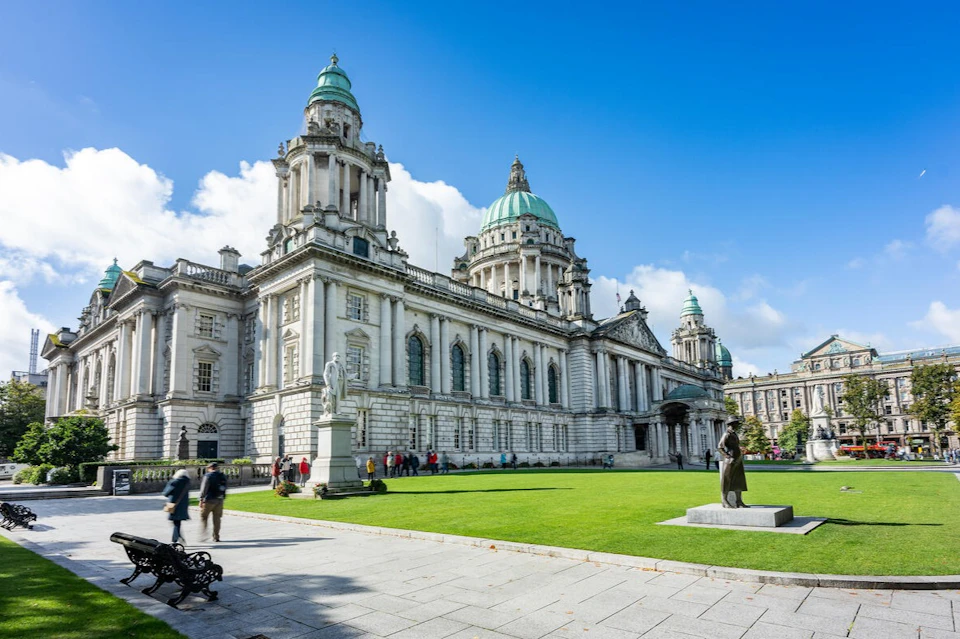 A grand white building with green domes, statues, and a manicured lawn under a blue sky with some clouds.