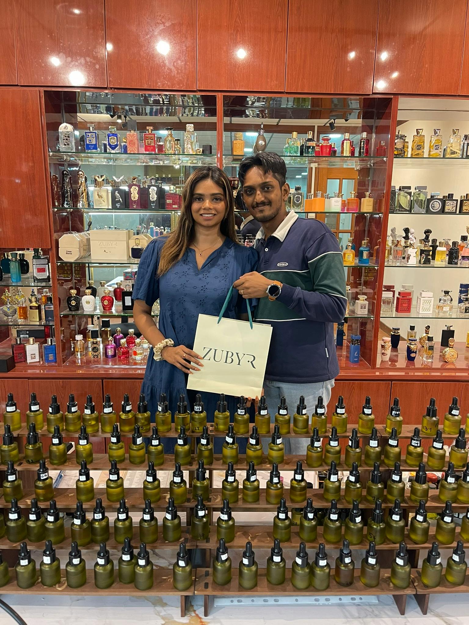 A man and woman stand in front of a display of perfume bottles. The woman holds a branded bag with the word 'Zubyr' on it.