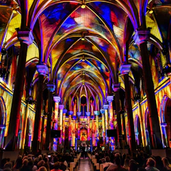 Interiören i en katedral, levande ljusprojektioner på valven och taket, med människor som sitter och tittar på displayen.