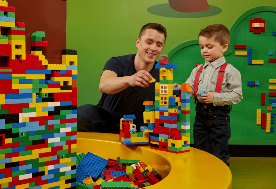 An adult and child happily build a colorful structure with LEGO bricks on a yellow table. Green background wall.
