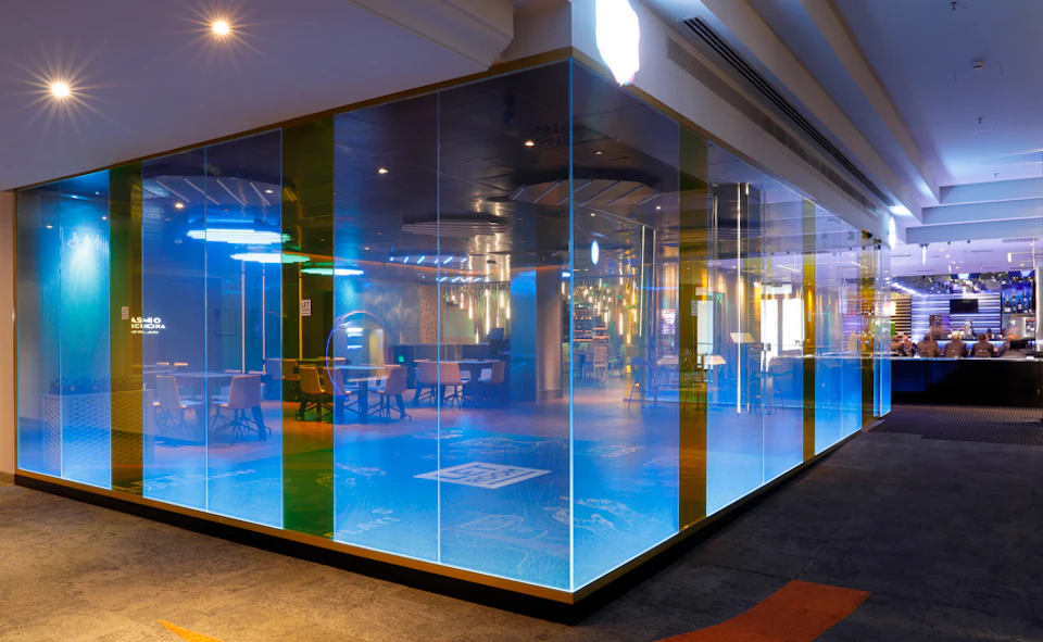 A modern, glass-walled room with colorful lighting, empty chairs, and tables inside, reflecting a sleek, contemporary design.