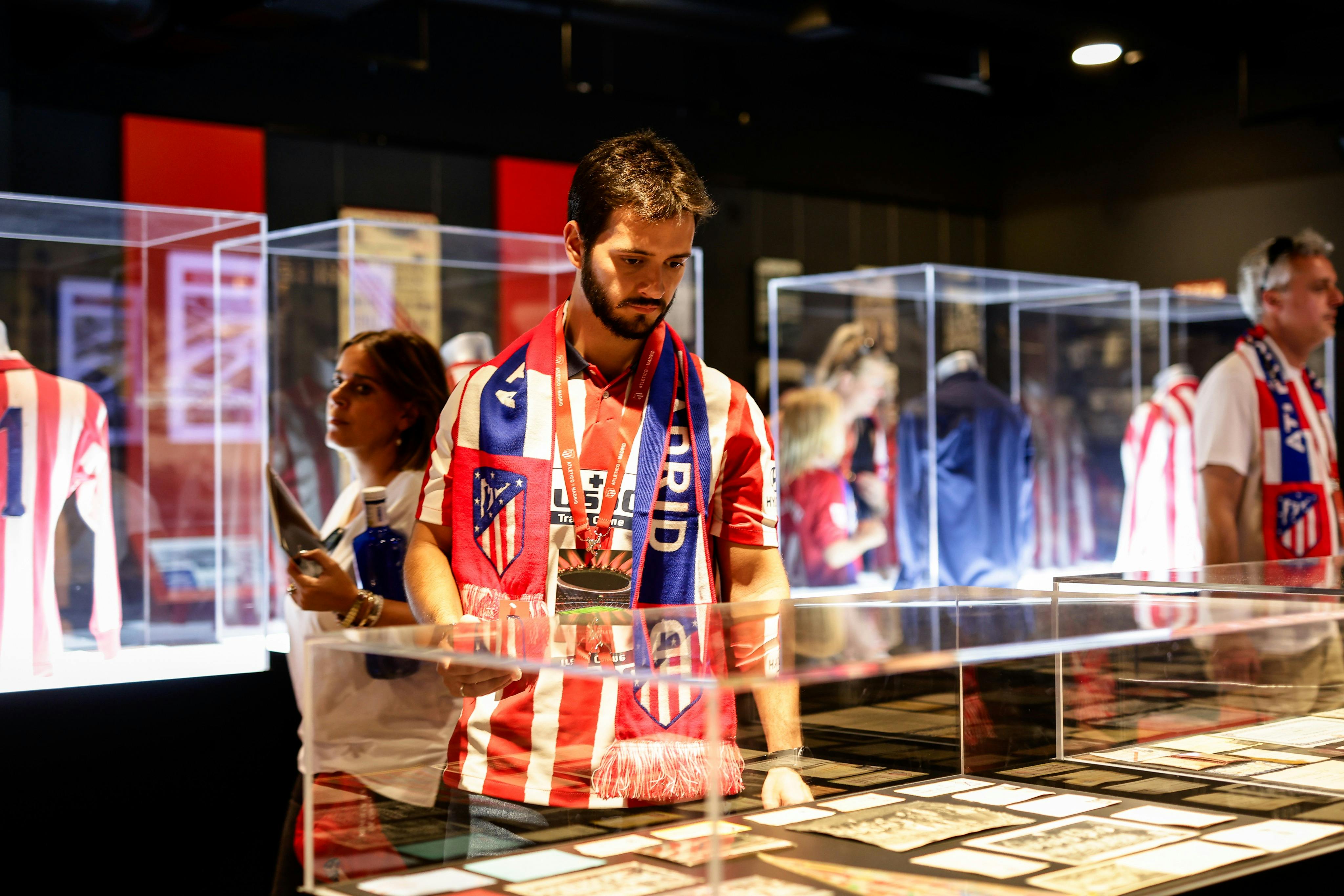 Een man in een gestreept shirt en sjaal bekijkt voorwerpen in een glazen vitrine, met een andere persoon in de buurt in een museum of tentoonstelling.