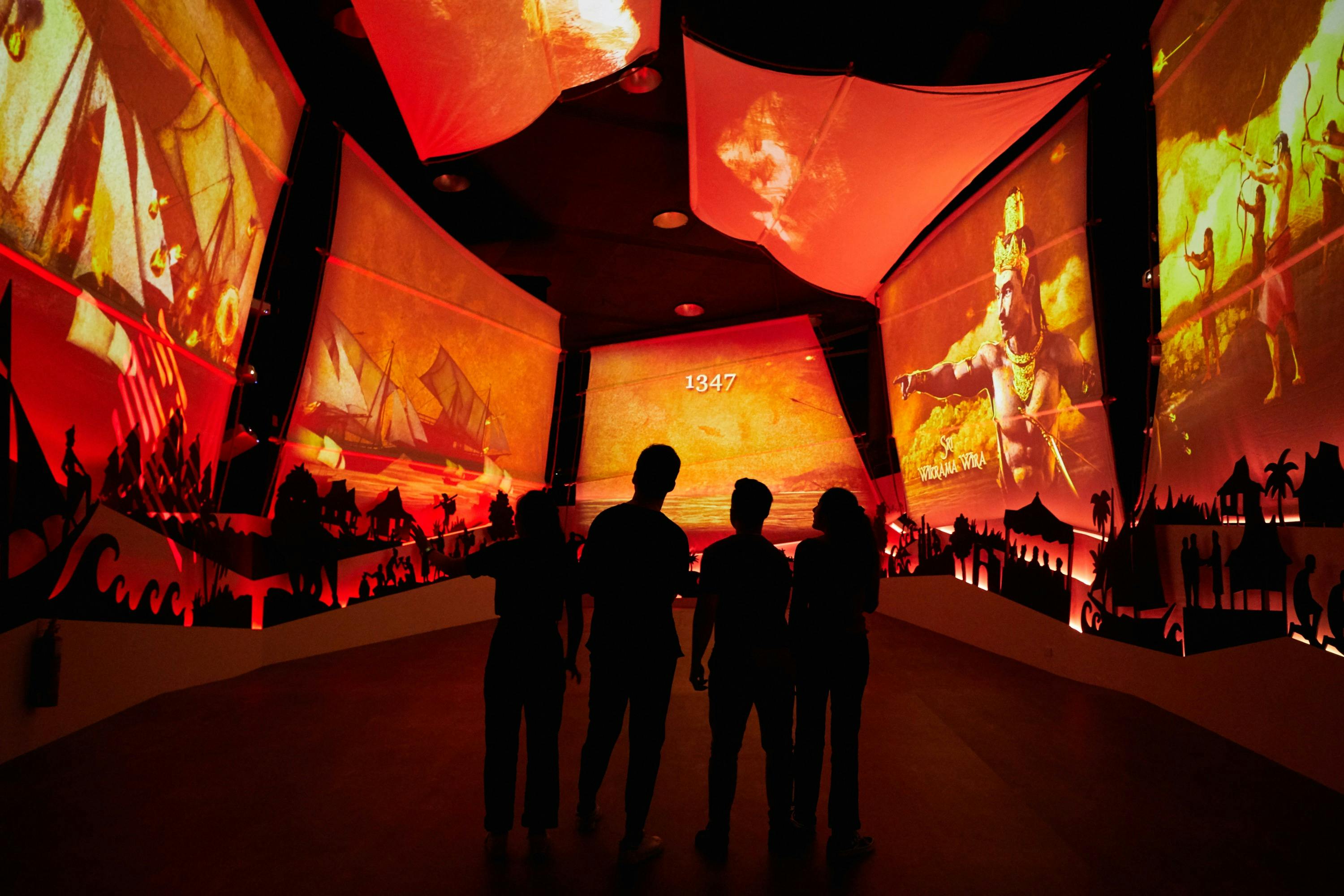Silhouettes of four people viewing an immersive, red-lit exhibit with historic imagery and projections on screens.