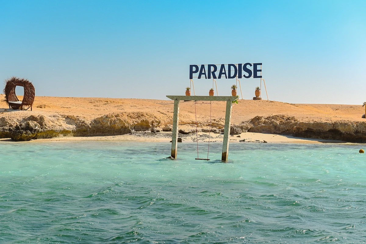 Wooden swing set in shallow turquoise water near a sandy shore, with a "PARADISE" sign and potted plants on top.