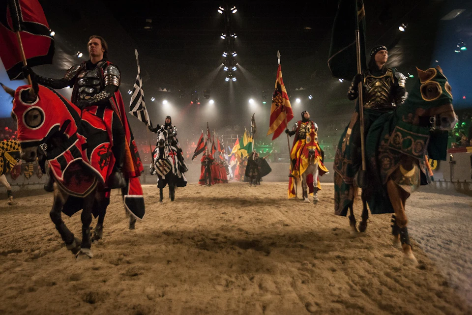 Knights on horseback in armor, holding banners, parade in an indoor arena with sand floor and audience in the background.