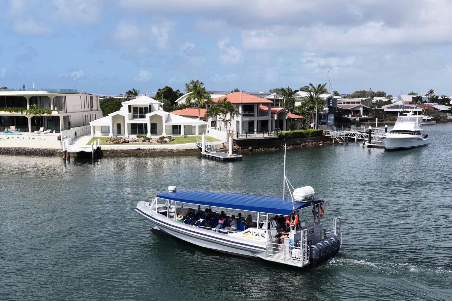 Een boot met een blauwe luifel die passagiers vervoert op een rustig water, met huizen aan de waterkant en palmbomen op de achtergrond.