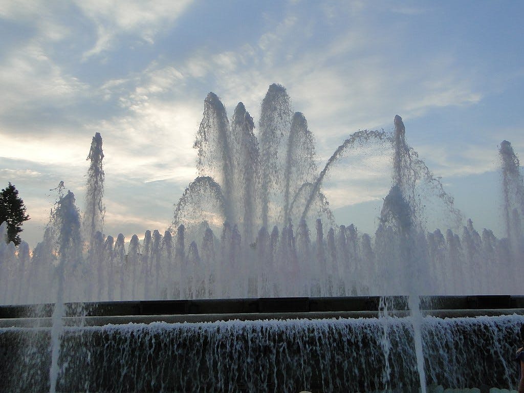 Fontaine avec de hauts jets d'eau sur fond de ciel nuageux, la lumière du soleil étant faiblement visible à travers l'eau. Arbres sur le côté gauche.