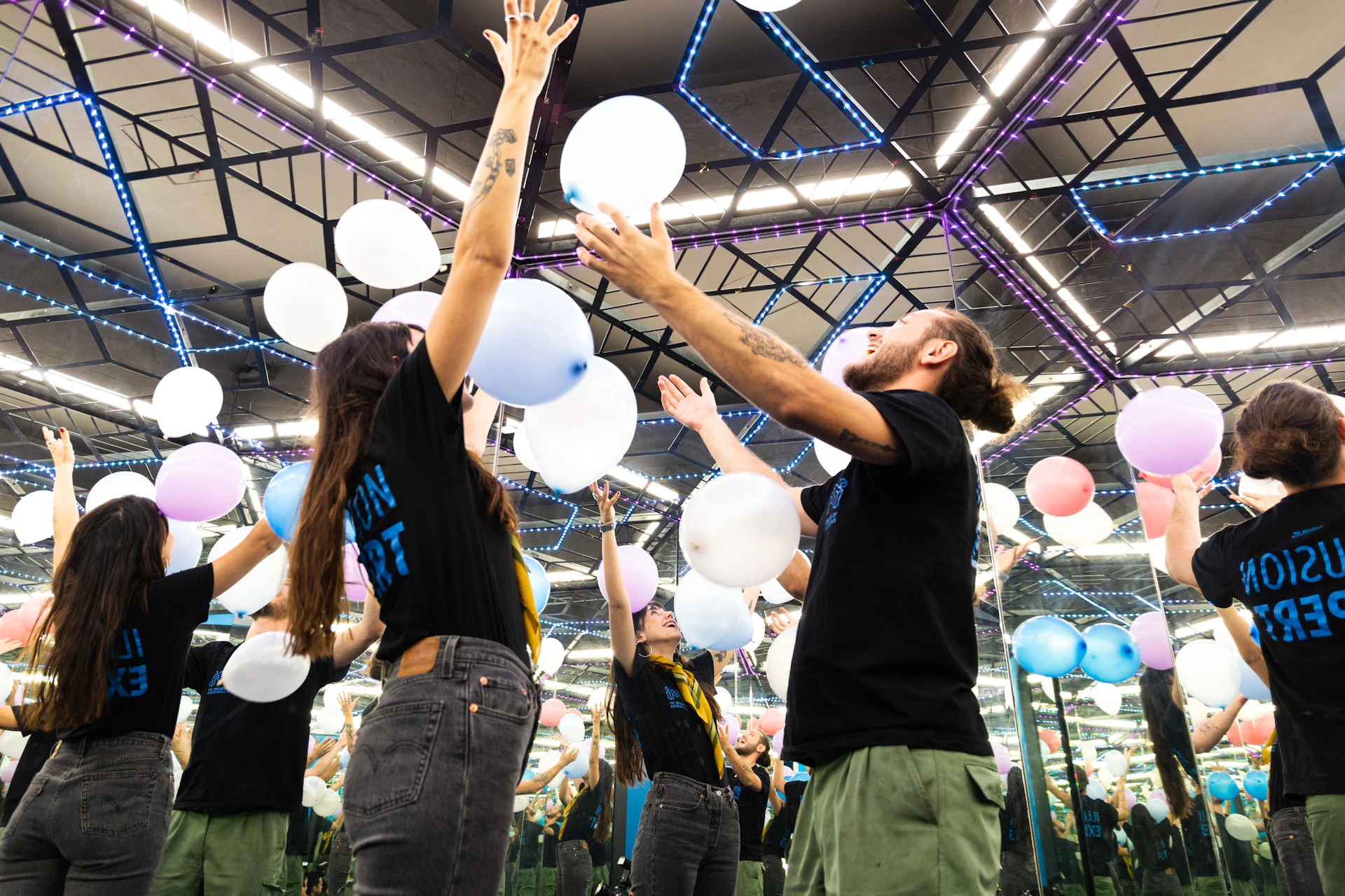 Two people in a room with mirrored walls and colourful lights toss white balloons into the air, creating a playful atmosphere.