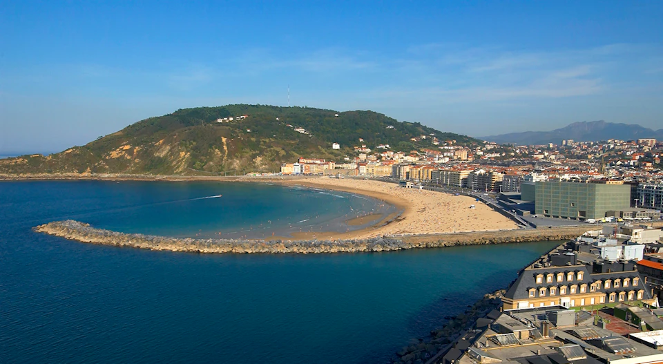 A coastal city with sandy beach, beachfront buildings, a breakwater, and surrounding green hills under a clear blue sky.