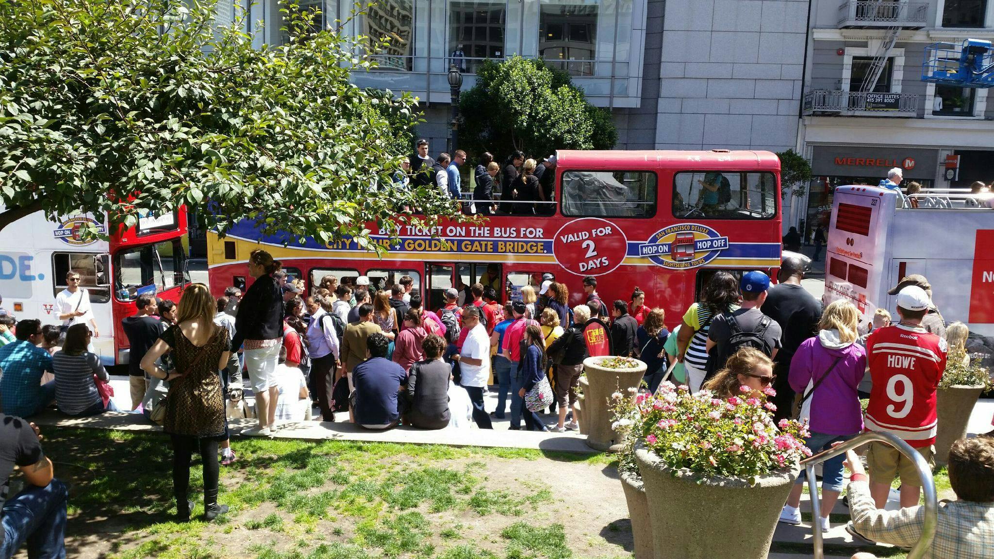 Une foule de personnes attend de monter à bord d'un bus touristique rouge à impériale près d'un bâtiment, avec un arbre et des pots de fleurs à proximité.