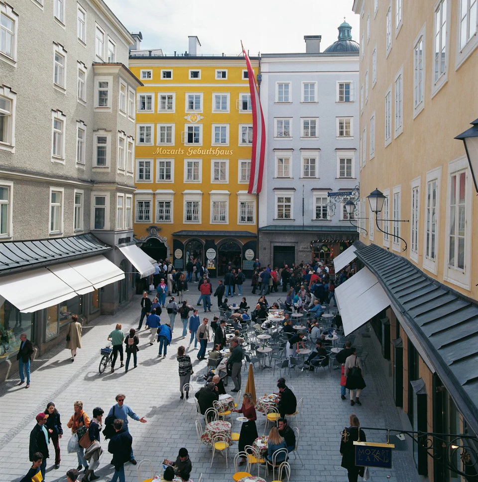 People walking and sitting at outdoor cafés in a European plaza, surrounded by yellow, gray, and white buildings.