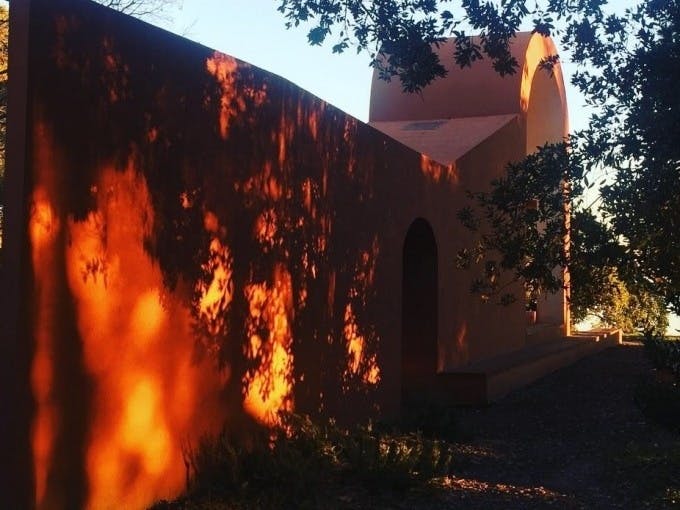 Солнечный свет отбрасывает тени на здание из оранжевой штукатурки с арочными дверными проемами, обрамленными листовыми ветвями.