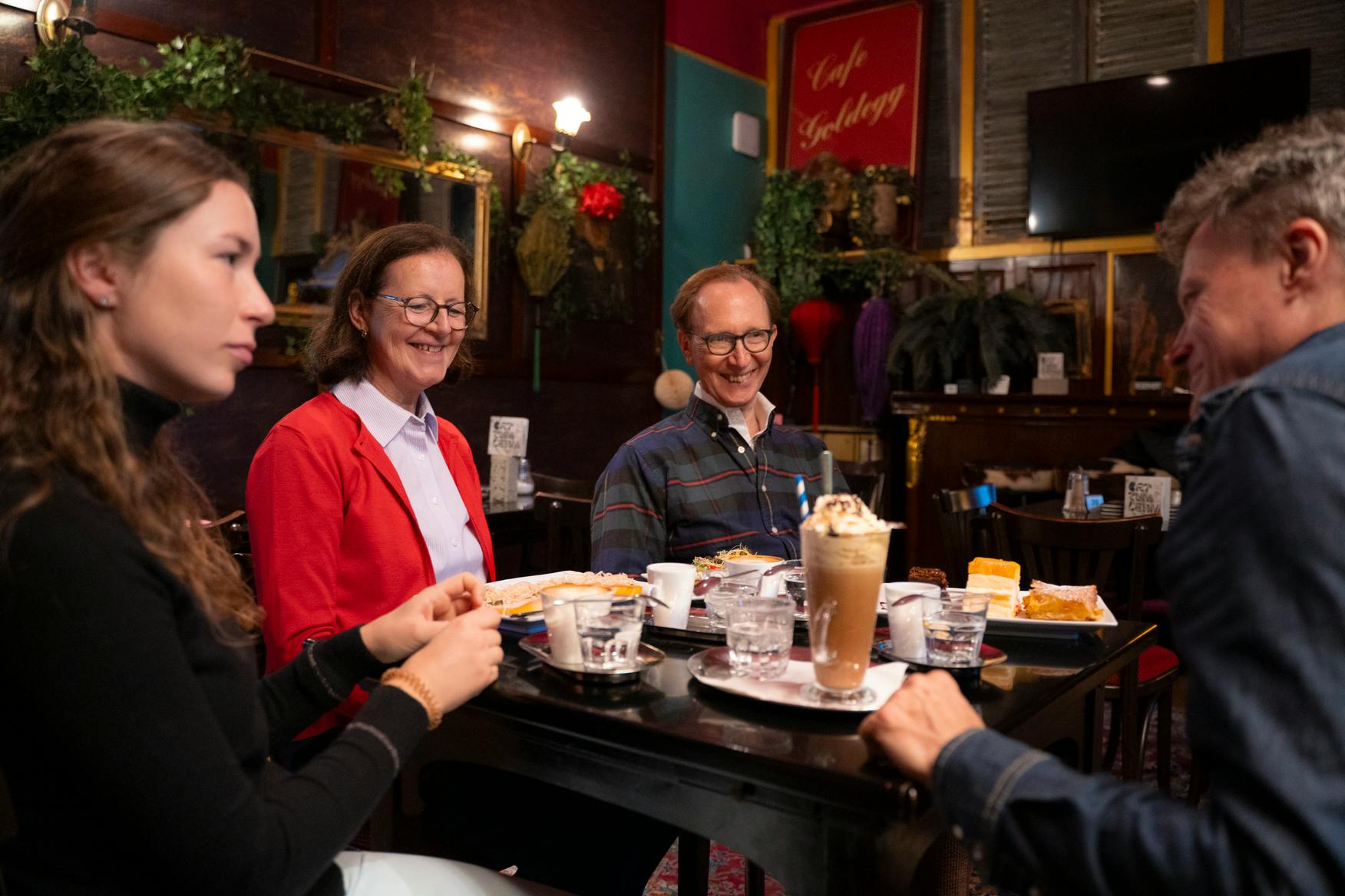 Três pessoas sentadas à mesa de um café com comida e bebidas, rodeadas por uma decoração festiva, a desfrutar do seu tempo juntos.