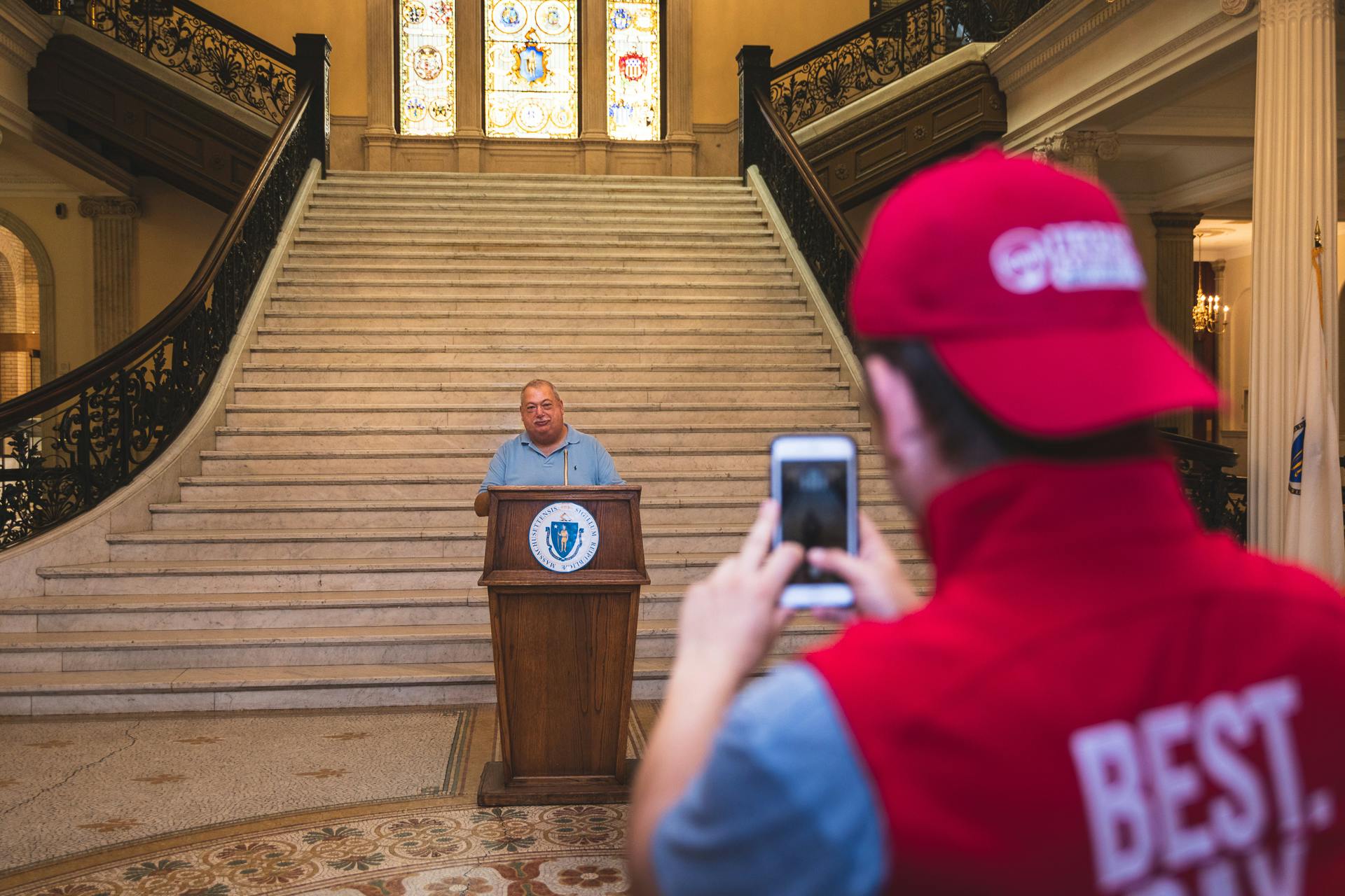 A person stands at a podium in front of a grand staircase, while another person in a red hat takes a photo with a smartphone.