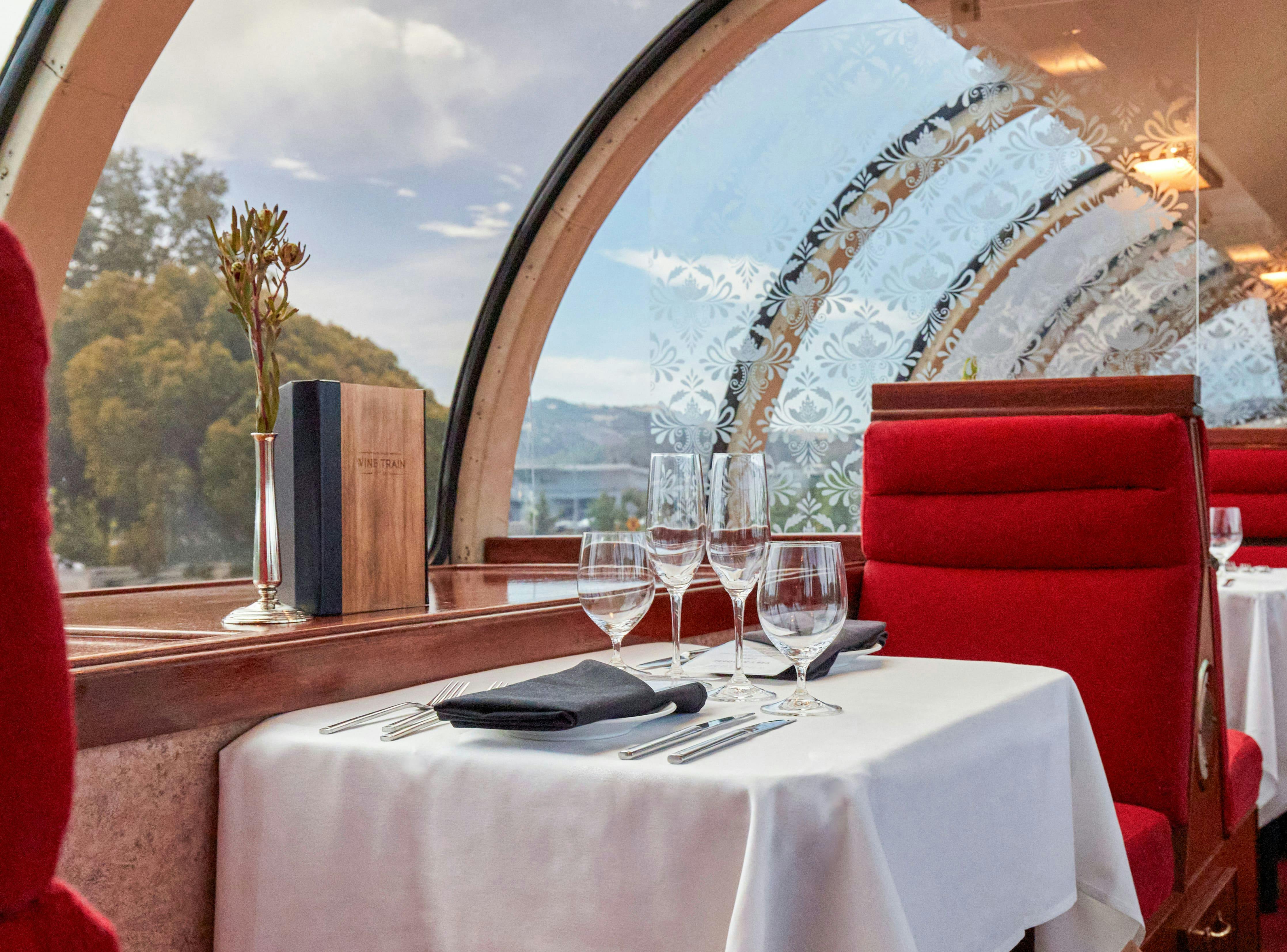 A table set for two with glassware and utensils in a scenic restaurant with large curved glass windows and red seating.