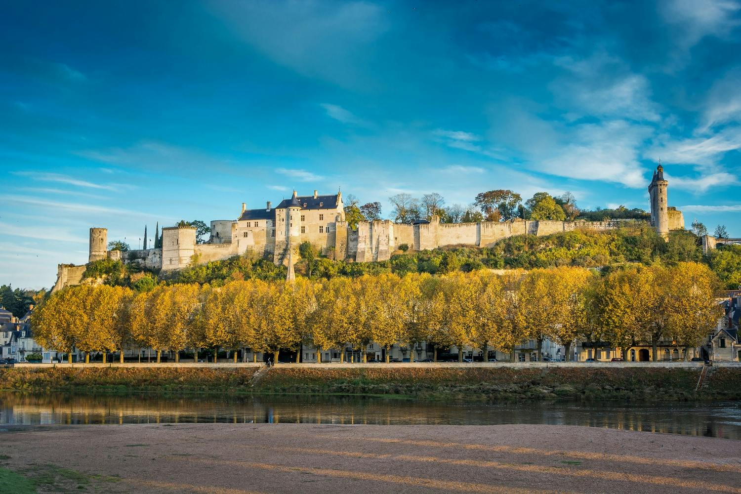 Un castello storico con alberi gialli autunnali in primo piano, sullo sfondo di un cielo blu e una riva del fiume in basso.