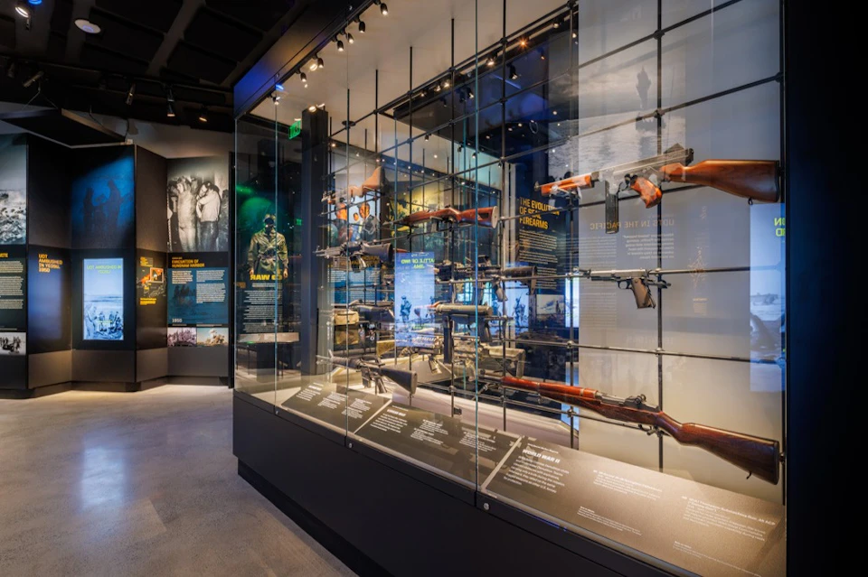 Museum display with firearms and historical artifacts in glass cases, accompanied by informational panels and lighting.