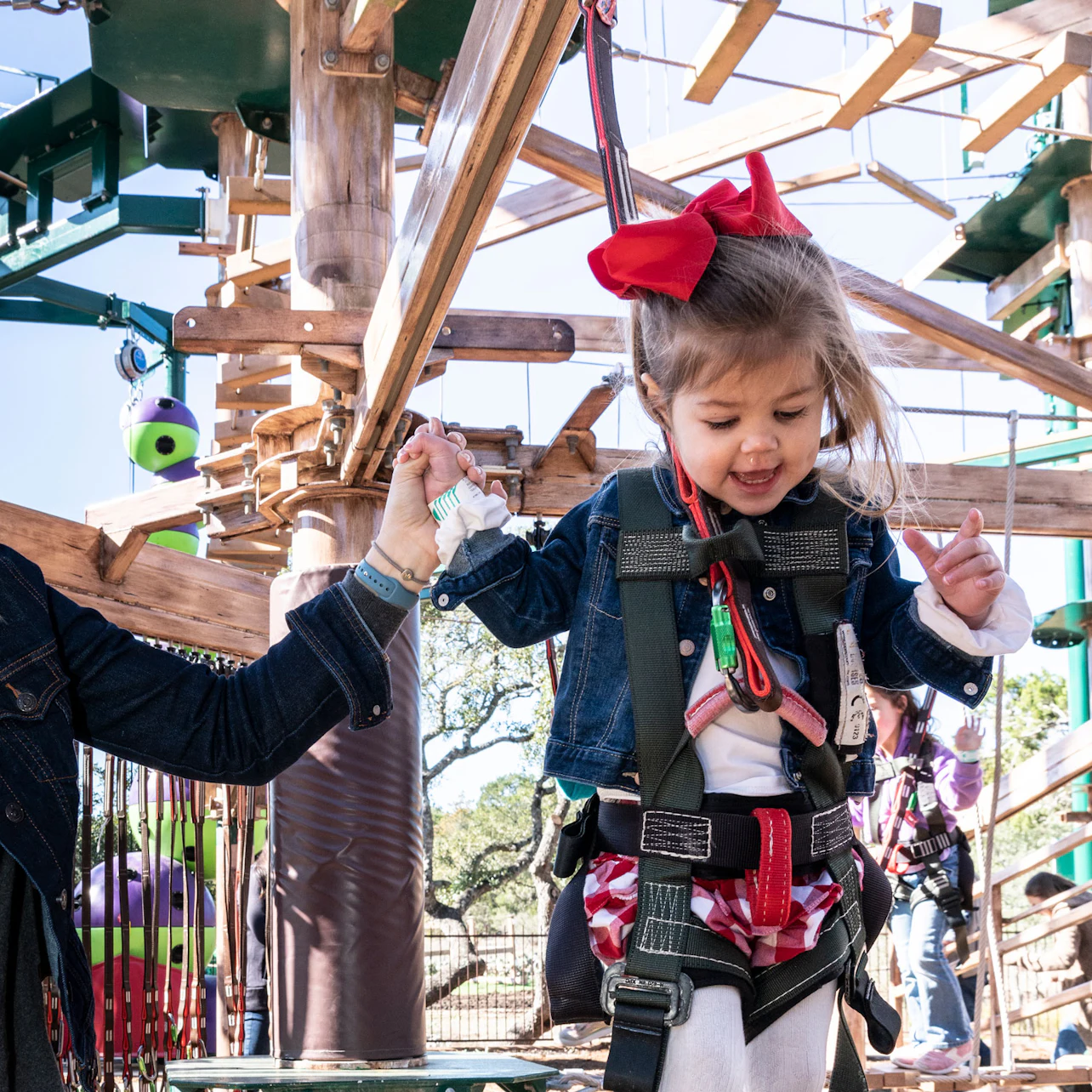 A young girl in a harness holds an adult’s hand while navigating a ropes course, smiling and wearing a red bow.