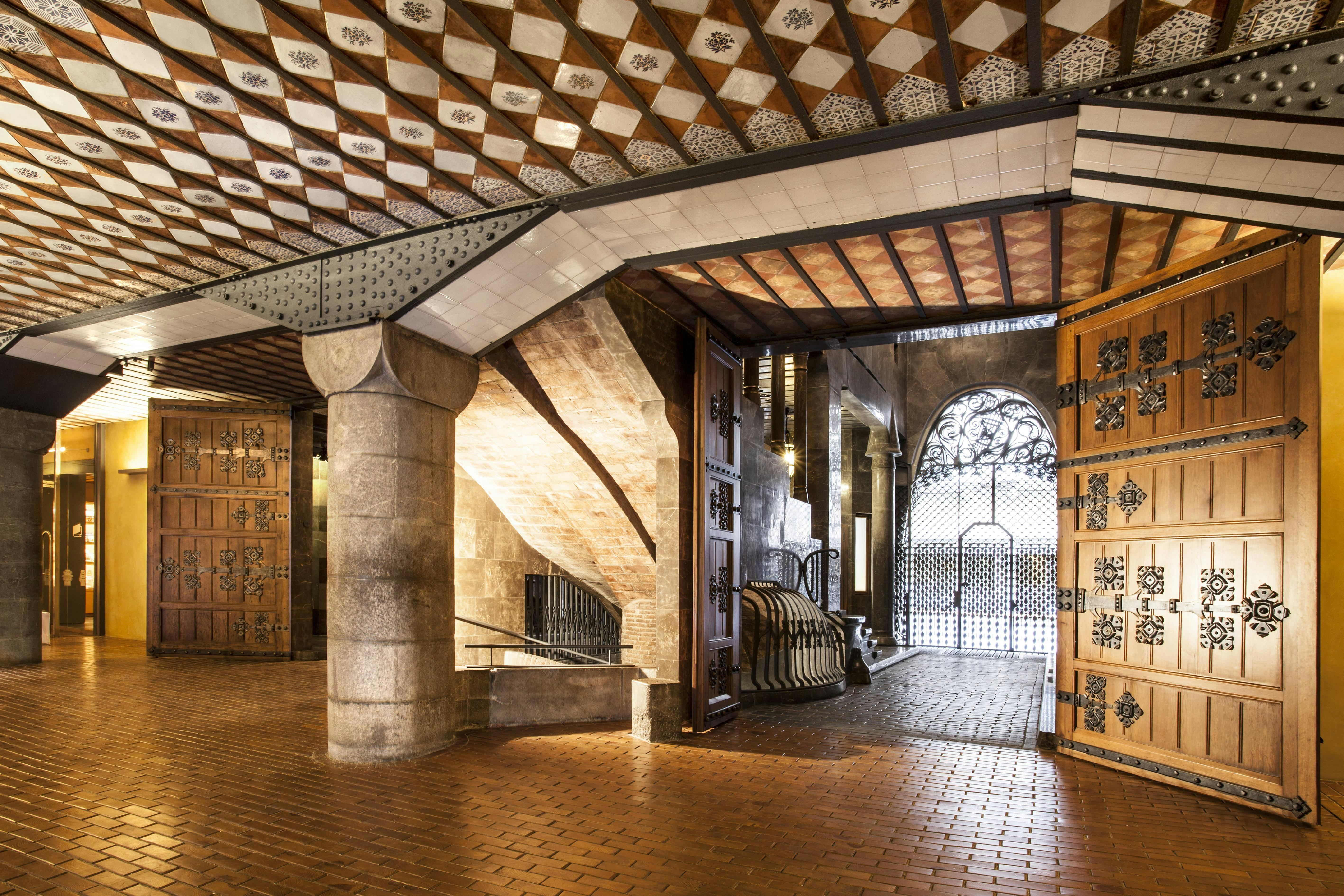 Interior of a building with ornate wood and metal doors, brick flooring, a curved stone staircase, and decorative ceiling tiles.
