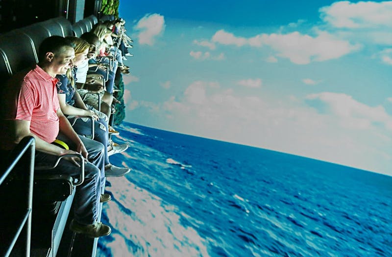 Люди, сидящие рядами на аттракционе, наклоняются влево, с проецируемым океаном и небесным фоном.