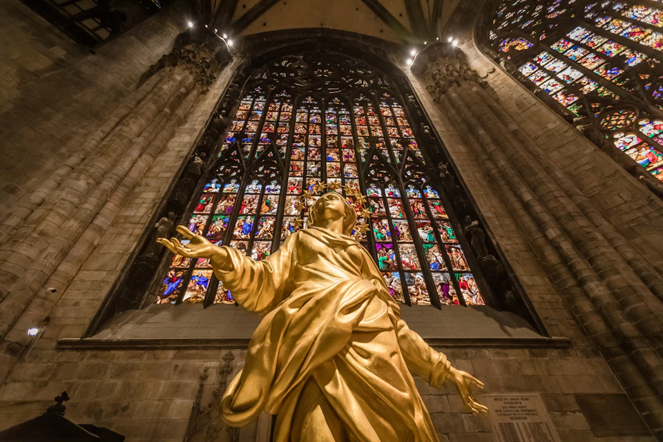 Golden statue in front of a large, ornate stained glass window within a cathedral, with intricate details and colorful patterns.
