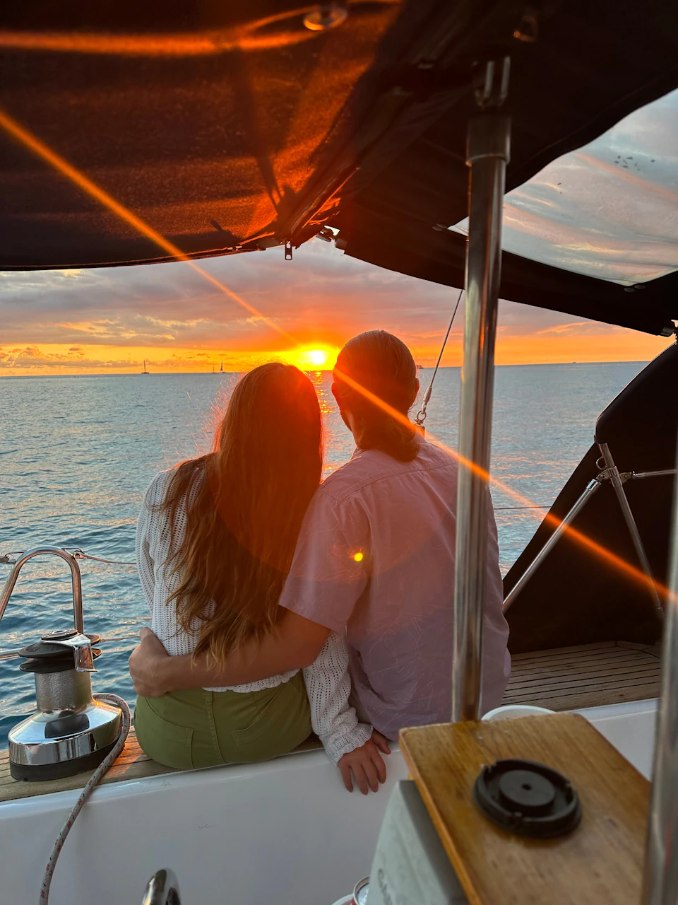 Two people seated on a boat at sunset, facing the ocean, with the sun casting an orange glow over the water.