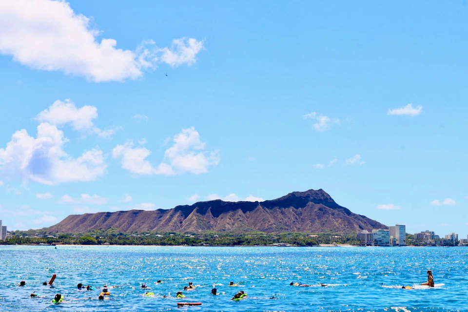 Swimmers and snorkelers in a blue ocean with a mountainous landscape and a clear, sunny sky in the background.