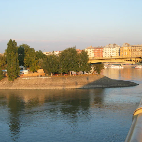 En flod med en liten båt, trädkantad strand, byggnader och en bro med flaggor i bakgrunden under solnedgången.