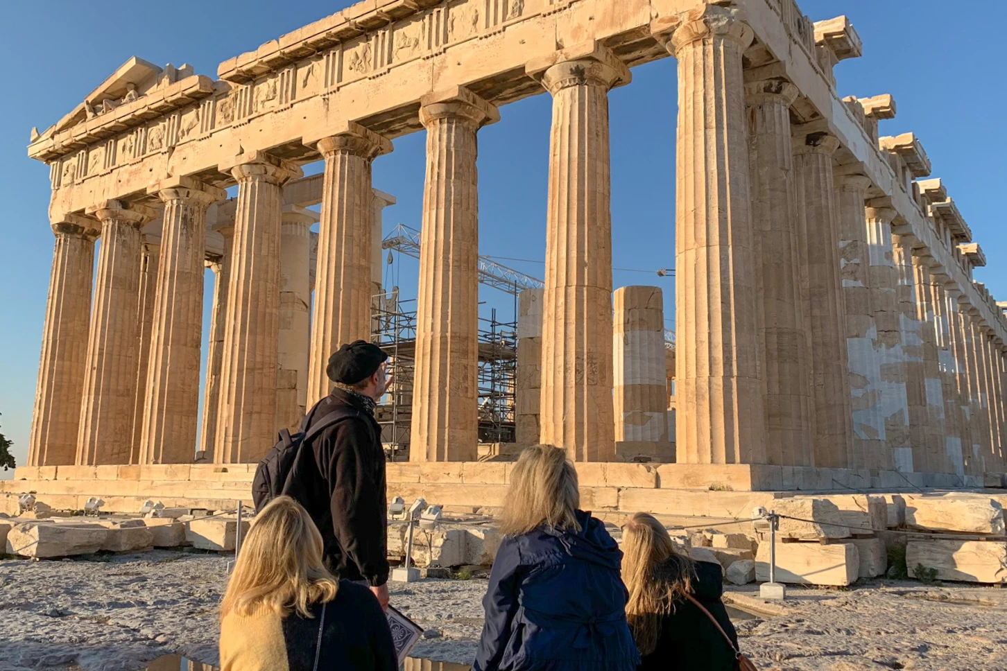 Four people observe the Parthenon, an ancient Greek temple with tall columns, under a clear blue sky. Scaffolding is visible inside.
