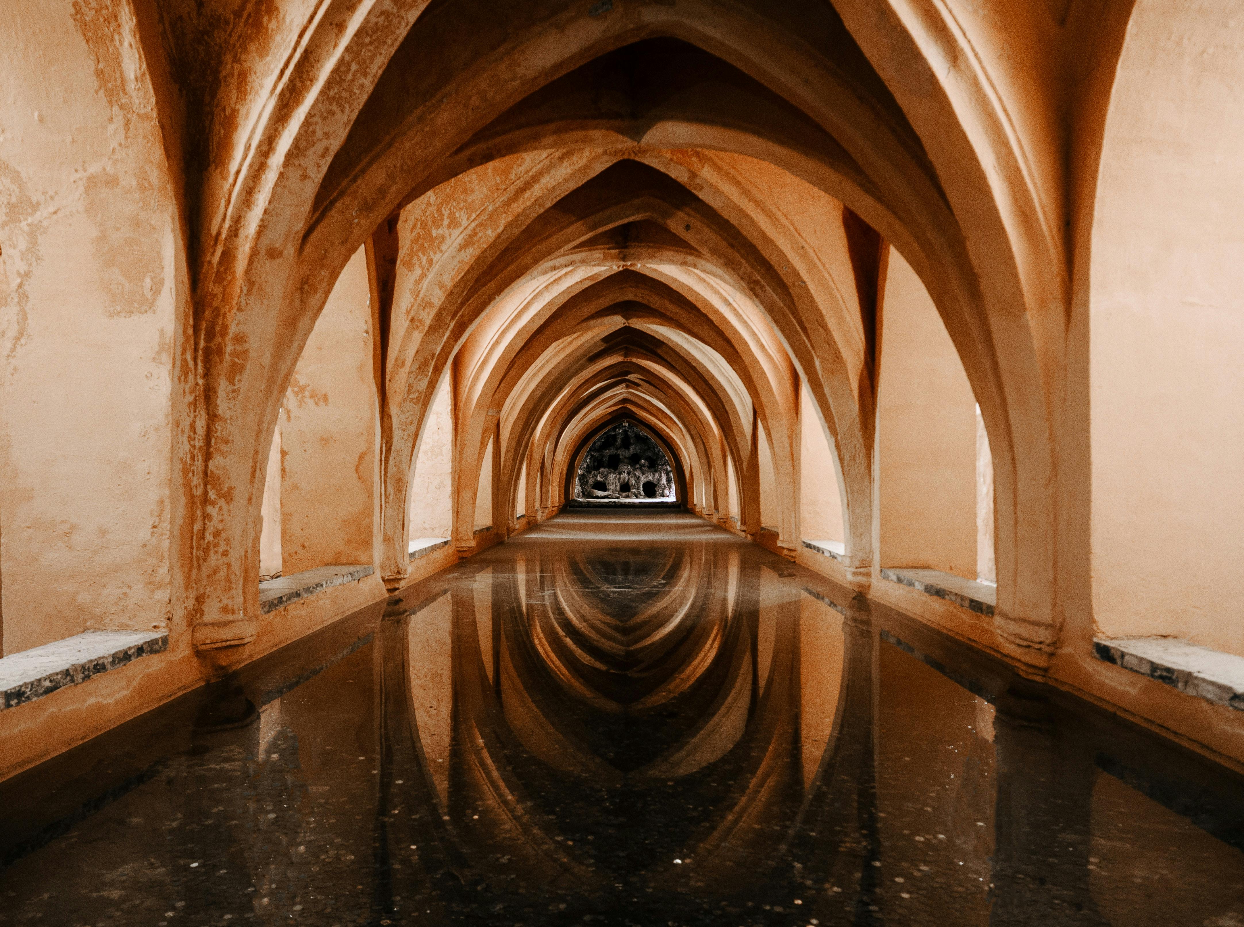 바닥에 반사된 물이 있는 아치형 석조 복도는 따뜻한 톤으로 빛나는 먼 아치형 개구부로 이어집니다.